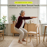 Frau ab 60 macht im Wohnzimmer eine Mini-Kniebeuge vor einem Holzstuhl, auf dem Sideboard steht die Übungskarte Mini-Kniebeuge – Hüfte, Gesäß, Beine