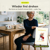 Frau ab 60 dreht sich am Esstisch im Sitzen zur Seite, die Übungskarte Oberkörper drehen steht im Kartenhalter