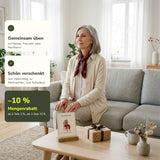 Frau ab 60 sitzt aufrecht auf dem Sofa, die Übungskarte Nacken + Schultern steht neben einem Geschenk auf dem Couchtisch, Mengenrabatt ab 2 Sets